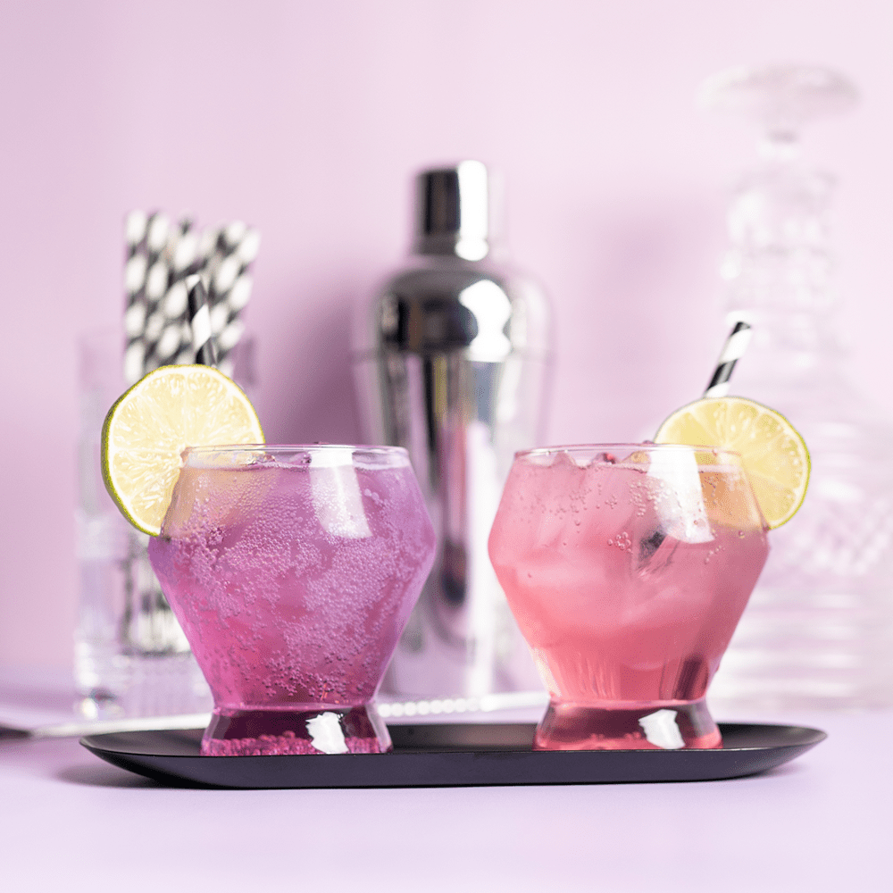 Pink and purple cocktails in whiskey glasses with lime slices on the rims and cocktail supplies in the background with soft studio lighting