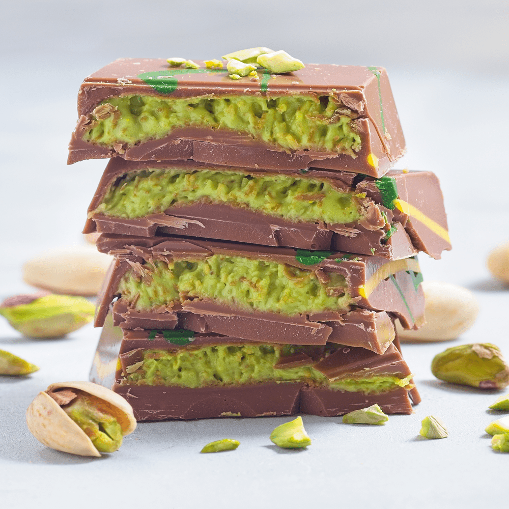 A stack of four broken chocolate bars filled with pistachio, photographed on a light gray surface with a few pistachios scattered around. The chocolate is dark brown and the filling is light green.