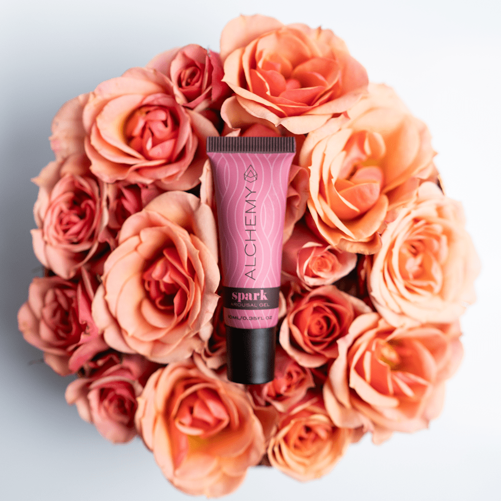 Pink tube of Alchemy Spark Female Arousal Gel surrounded by peach roses on a light gray background