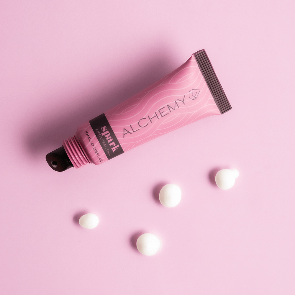 Alchemy Spark Female Arousal Gel tube on a pink background with light liquid droplets