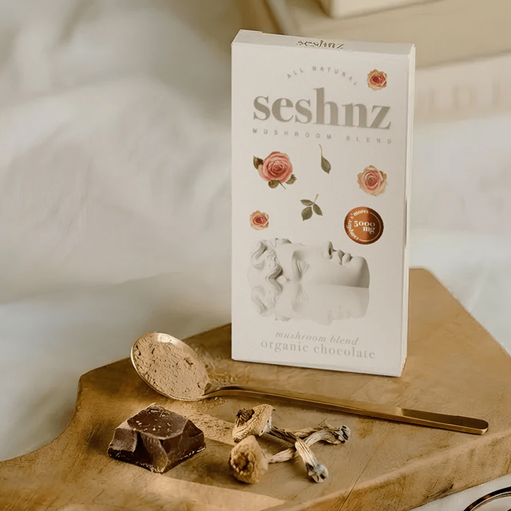 A box of Seshnz Mushroom Blend organic chocolate on a wooden board with a gold spoon, chocolate pieces, and dried mushrooms against a soft white fabric background