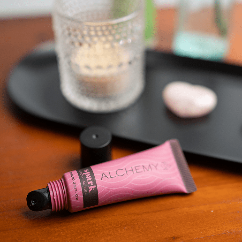 Pink tube of Alchemy Spark Female Arousal Gel on a wooden surface with a blurred candle in the background