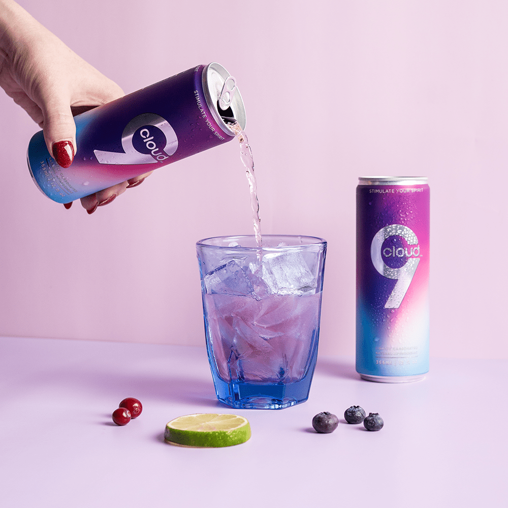 Person's hand pouring a can of Cloud 9 Aphrodisiac Drink into a glass on a light purple surface with cherries, blueberries, and a lime slice in the foreground