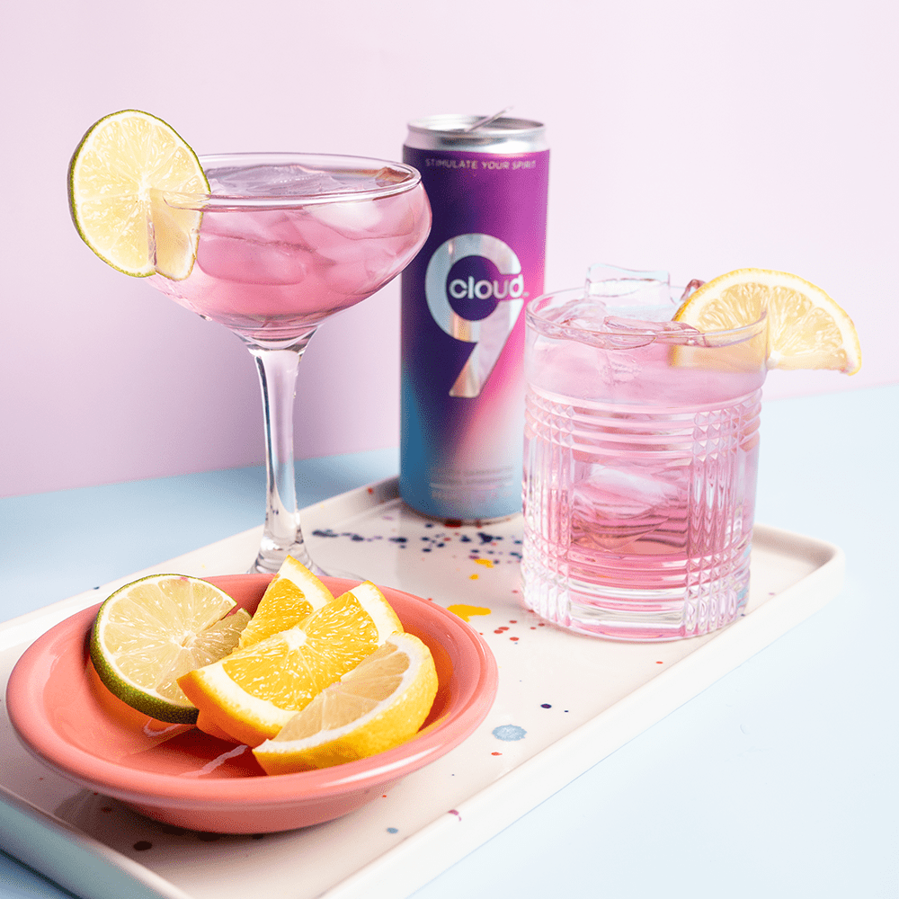 Pink cocktails in cocktail glasses with lime and orange slices and a can of Cloud 9 Aphrodisiac Drink in the background on a light surface with soft lighting