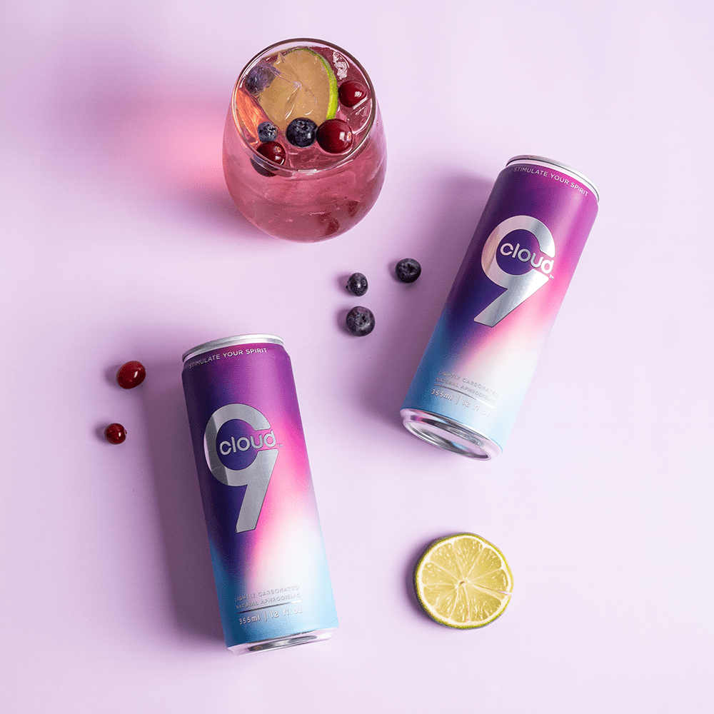 Two Cloud 9 Aphrodisiac Drink cans with a glass of a pink drink and fruit on a pink background with soft lighting