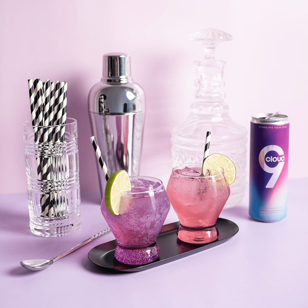 Cloud 9 Aphrodisiac Drink: purple aluminum can with martini shaker, crystal glass container, striped straws, and pink and purple cocktails in whiskey glasses set up in a studio environment with soft lighting