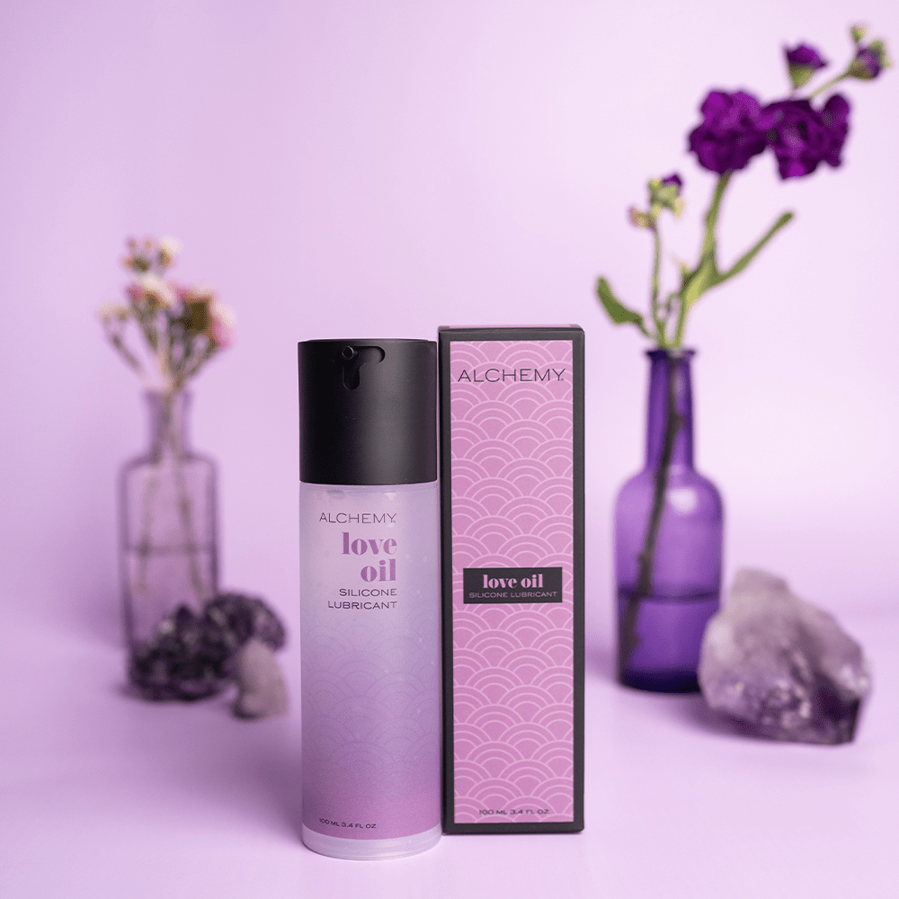 Alchemy Love Oil Silicone Personal Lubricant bottle and box with plants in vases and purple background