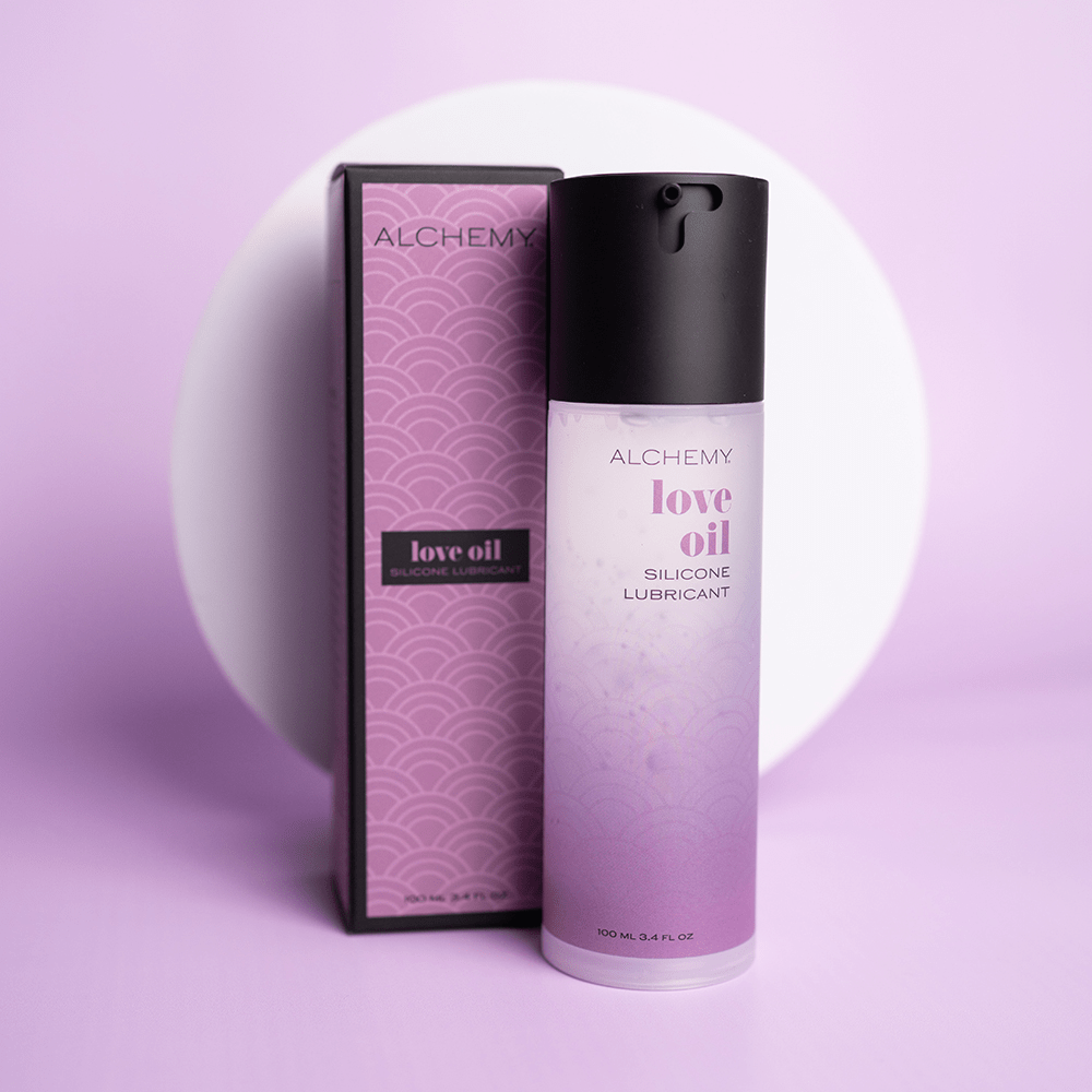 Alchemy Love Oil Silicone Personal Lubricant bottle and box on purple surface with a white circular shape in the background