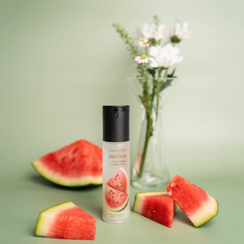 Alchemy Indulge – Watermelon Flavored Personal Lubricant bottle on a green surface with watermelon slices and flowers in a vase