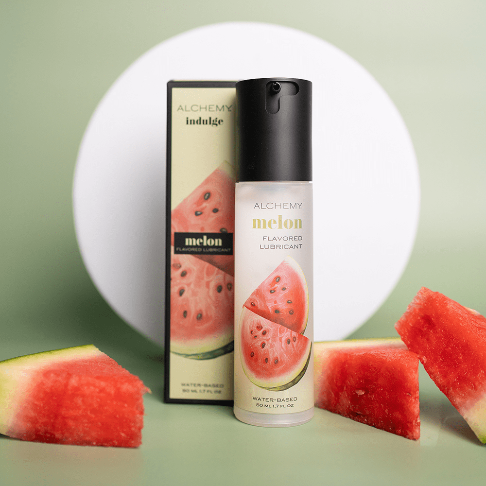 Alchemy Indulge – Watermelon Flavored Personal Lubricant bottle and box with watermelon slices on a green surface with a white circular shape in the background
