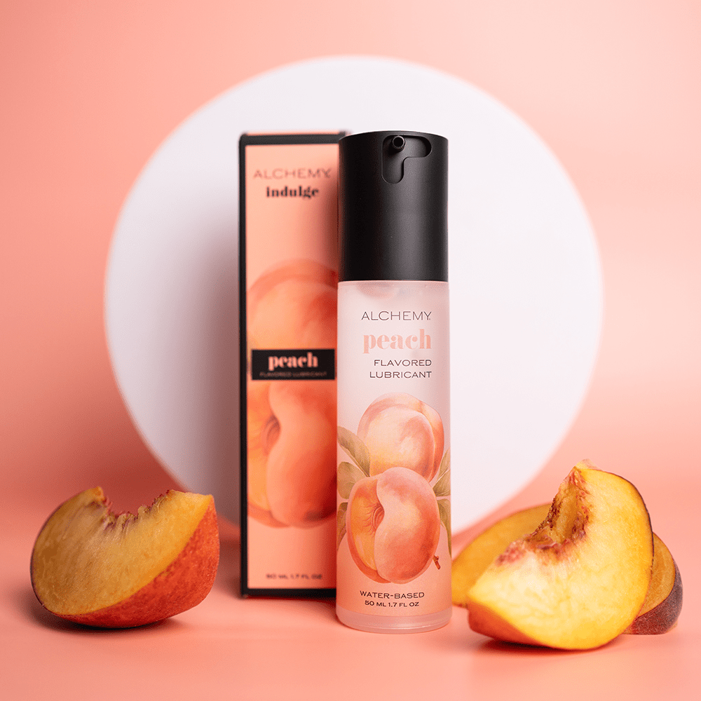 Alchemy Indulge – Peach Flavored Personal Lubricant bottle and box with peach slices on a peach-colored surface with a white circular shape in the background