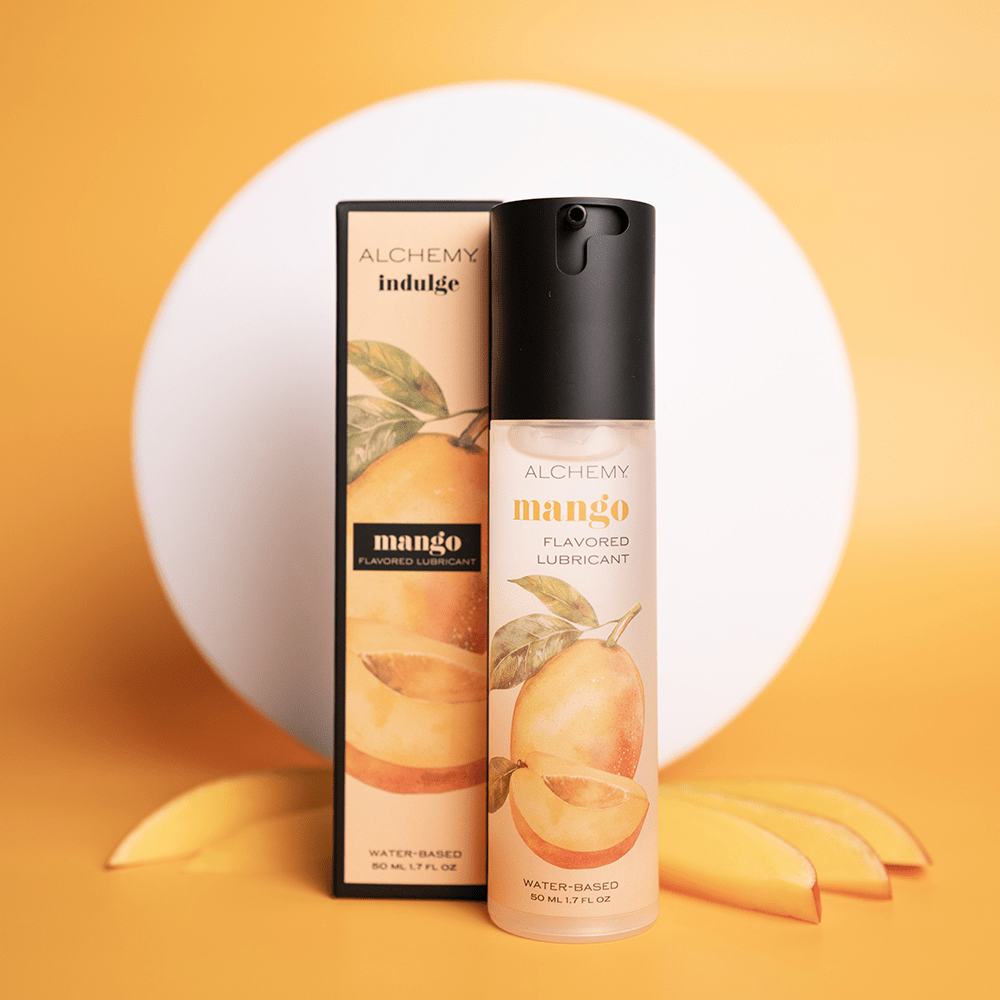 Alchemy Indulge – Mango Flavored Personal Lubricant bottle and box with mango slices on a yellow surface with a white circular shape in the background