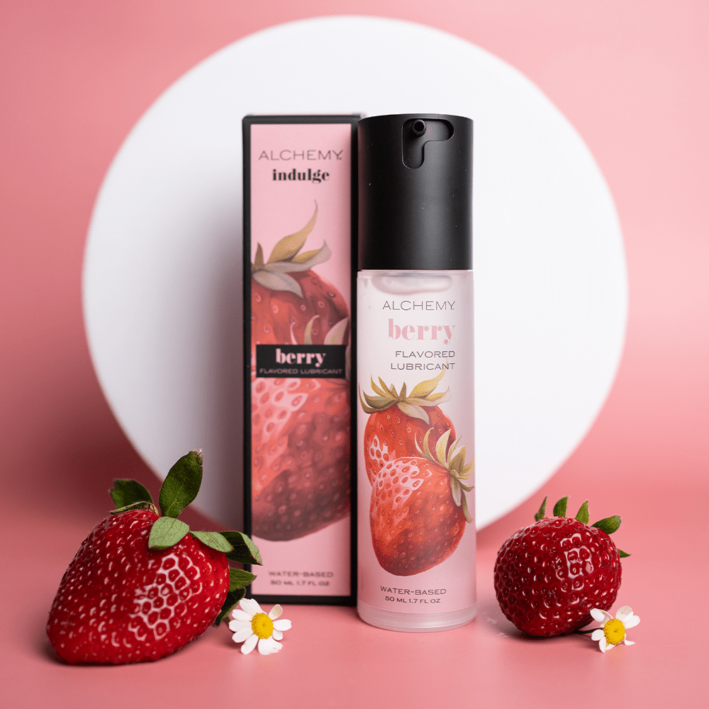 Alchemy Indulge – Strawberry Flavored Personal Lubricant bottle and box with fresh strawberries and a white shape in the background on a pink surface