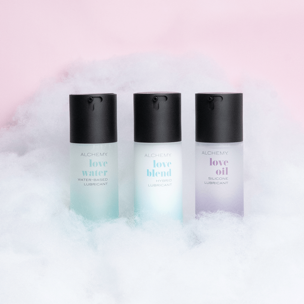 Amazing Intimate Essentials Alchemy Discovery Kit: Alchemy Love Water, Love Blend, and Love Oil personal lubricant bottles on a fluffy cloud texture against a pink background