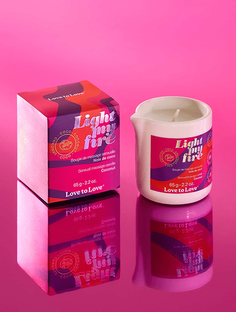 Love to Love Cosmetics Light My Fire Sensual Massage Candle and box on a mirror surface with a pink background
