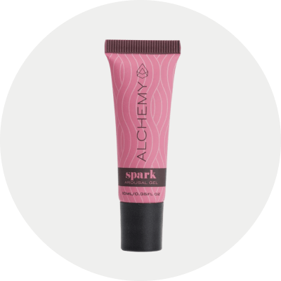 A pink and black squeeze tube of Alchemy Spark Female Arousal Gel, centered on a light gray circular background