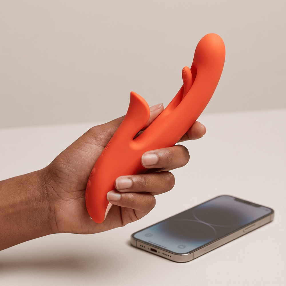 Person's hand holding Honey Play Box Tressa App-Controlled Tapping Rabbit Vibrator next to a smartphone with a beige background and studio lighting