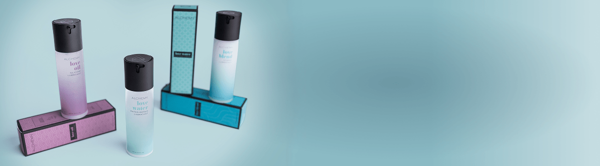 Three bottles of Alchemy personal lubricants and packaging on a light blue studio background