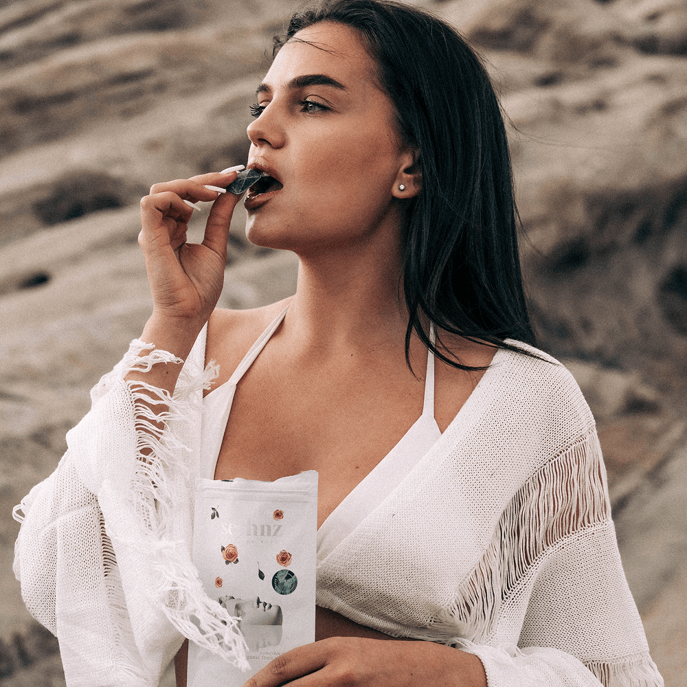 Woman in a white outfit with fringe holding a Seshnz functional mushroom chocolate package and eating a chocolate bit against a desert background